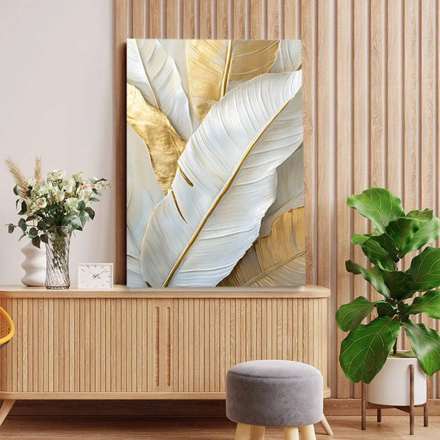 Large vertical wall art featuring oversized white and gold tropical leaves, displayed above a light wood console in a styled interior with a plant, vase, stool, and wood-paneled wall.