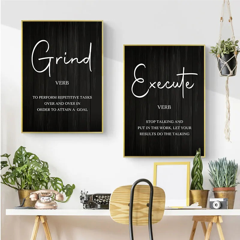 Two framed motivational wall art prints hang on a white wall above a styled desk with plants, a typewriter, and a chair. The left print says 'Grind' and the right print says 'Execute' in white script and serif text on a black wood-grain background with thin gold frames.