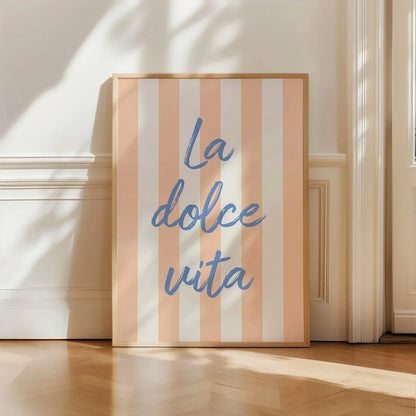 A framed vertical art print with vertical peach and white stripes and the blue script text 'La dolce vita', leaning against a cream wall on a light wood floor.